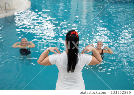 Instructor leading aquatic fitness training for group senior women in a swimming pool. Concept of health, exercise, wellness, water therapy, and active aging lifestyle Instructor leading aquatic fitness training for group senior women in a swimming pool. Concept of health, exercise, wellness, water therapy, and active aging lifestyle 133352889