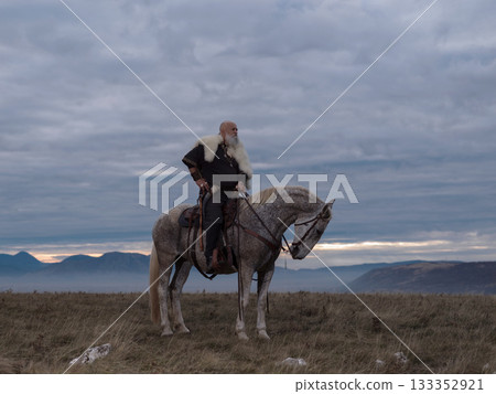 A majestic Viking chieftain on a dappled grey horse, clad in a fur cloak, surveys a desolate, mountainous landscape under a dramatic cloudy sky, exuding power and presence. 133352921