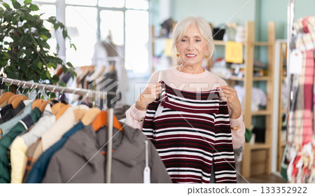 European elderly female buyer standing in clothing market and carefully examines selection of warm sweaters for winter season European elderly female buyer standing in clothing market and carefully examines selection of warm sweaters for winter season 133352922