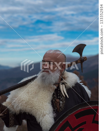 Bearded Viking warrior in fur cloak holding red shield and axe outdoors. Medieval Nordic fighter in traditional armor Bearded Viking warrior in fur cloak holding red shield and axe outdoors. Medieval Nordic fighter in traditional armor 133352934