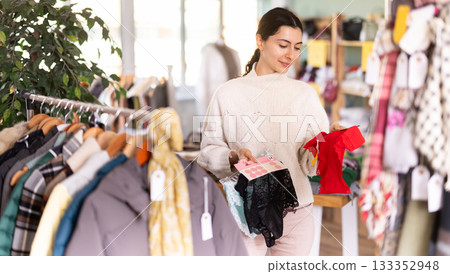 Happy young girl customer dressing in ordinary clothes choosing underwear and holding panties in hands in store Happy young girl customer dressing in ordinary clothes choosing underwear and holding panties in hands in store 133352948
