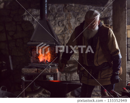 A Viking blacksmith with a white beard and braided hair works intently at a forge, hammering glowing metal on an anvil, with flames flickering in the background of a stone workshop. 133352949