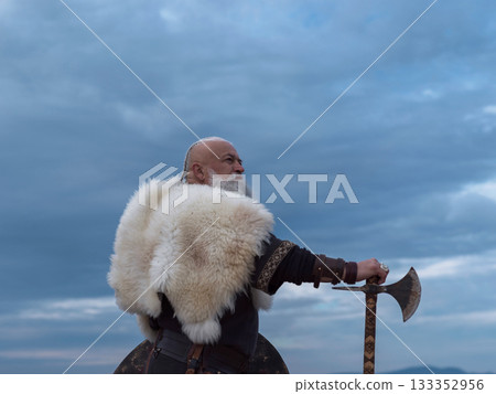 Viking warrior with axe standing on mountain cliff, wearing fur cloak and leather armor, overlooking dramatic landscape. Historical Nordic fighter 133352956