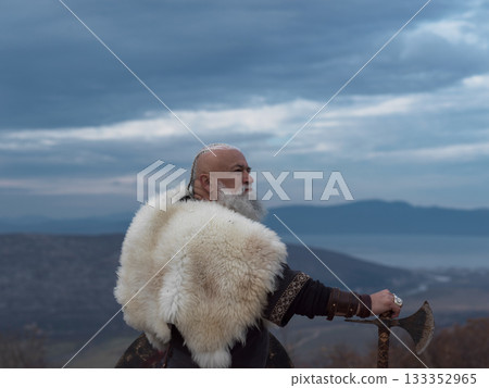 Viking warrior with axe standing on mountain cliff, wearing fur cloak and leather armor, overlooking dramatic landscape. Historical Nordic fighter Viking warrior with axe standing on mountain cliff, wearing fur cloak and leather armor, overlooking dramatic landscape. Historical Nordic fighter 133352965