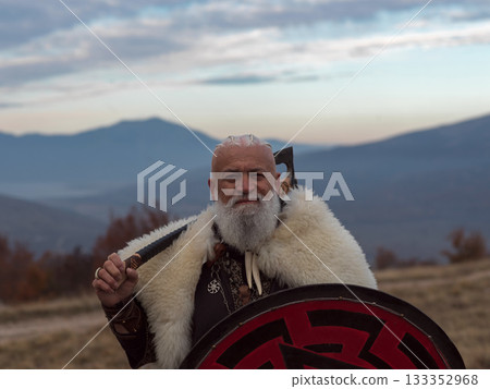 Bearded Viking warrior in fur cloak holding red shield and axe outdoors. Medieval Nordic fighter in traditional armor 133352968