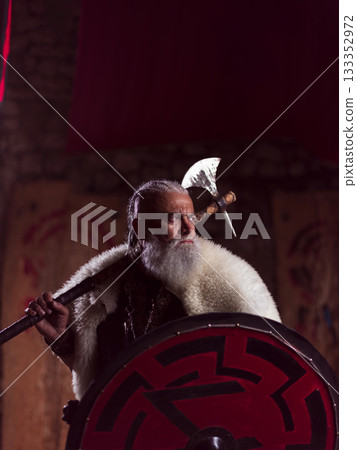 A resolute Viking chieftain, with a white beard and fur cloak, stands holding a formidable axe over his shoulder and a red and black round shield, embodying strength and ancient resolve. 133352972