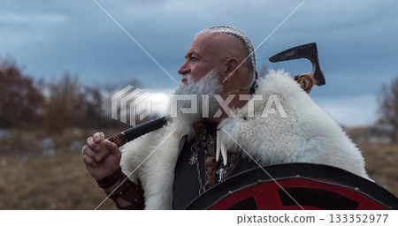 Bearded Viking warrior in fur cloak holding red shield and axe outdoors. Medieval Nordic fighter in traditional armor 133352977