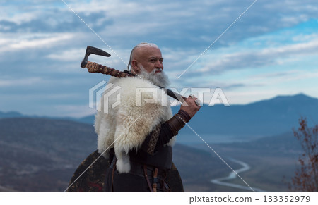 Viking warrior with axe standing on mountain cliff, wearing fur cloak and leather armor, overlooking dramatic landscape. Historical Nordic fighter 133352979