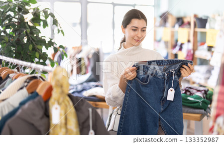 Happy pretty young girl client carefully choosing stylish jeans trousers in clothing store. Winter sales concept Happy pretty young girl client carefully choosing stylish jeans trousers in clothing store. Winter sales concept 133352987