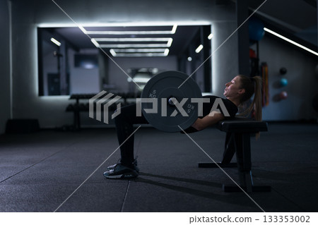 Strong woman performs hip thrusts with weight in modern gym during evening workout session 133353002