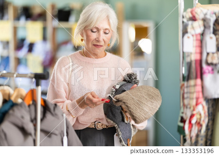 Elderly woman carefully selects warm hat in clothing store Elderly woman carefully selects warm hat in clothing store 133353196
