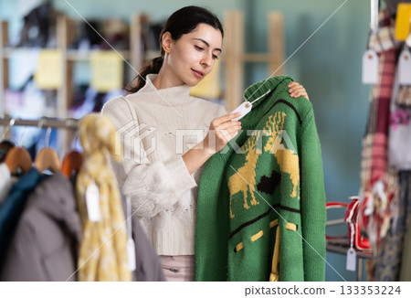 Interested young girl buyer standing in clothing market and carefully examines selection of sweaters on sale of winter clothing collection 133353224