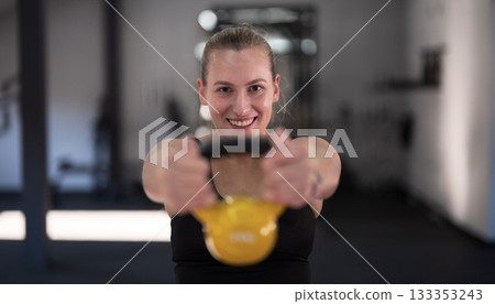 Woman exercises with yellow kettlebell in gym during daytime, showcasing strength and fitness routine 133353243
