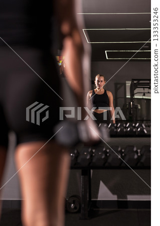 Woman exercising in gym while looking in mirror during strength training session in modern fitness center Woman exercising in gym while looking in mirror during strength training session in modern fitness center 133353246
