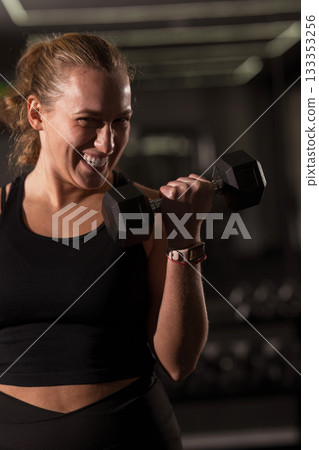 Young woman happily lifting dumbbell in gym during workout session in the evening 133353256