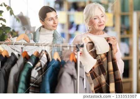 Family shopping concept. Two happy women choosing coat or jacket for cold season in clothing shop with large assortment Family shopping concept. Two happy women choosing coat or jacket for cold season in clothing shop with large assortment 133353307