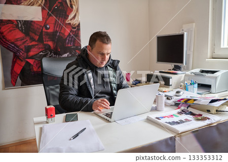 Man working on laptop at cluttered desk in office, surrounded by paperwork and office supplies, creating a busy work environment, indoor scene. Man working on laptop at cluttered desk in office, surrounded by paperwork and office supplies, creating a busy work environment, indoor scene. 133353312