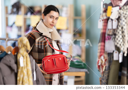 Sale of winter clothing collection. Positive young girl standing against background of shelves with clothes in store and choosing bag Sale of winter clothing collection. Positive young girl standing against background of shelves with clothes in store and choosing bag 133353344