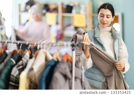 Focused young girl client selecting trousers in clothing shop. Winter clothing collection 133353425