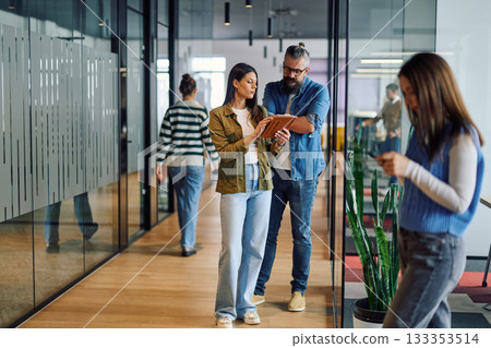 Two business professionals having a focused discussion while reviewing information on a tablet in a bright modern office hallway, symbolizing teamwork, communication, and digital collaboration. 133353514