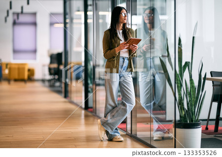 Professional business woman standing in modern office corridor, holding digital tablet and smiling confidently, representing leadership, technology, and success in a creative business environment. 133353526