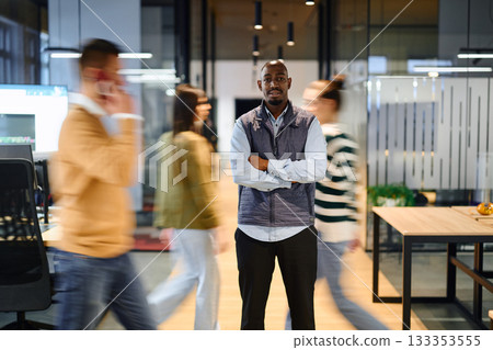 Confident african american businessman standing with crossed arms in the center of a busy office hallway while colleagues move around him, symbolizing leadership, focus, and stability in a dynamic 133353555
