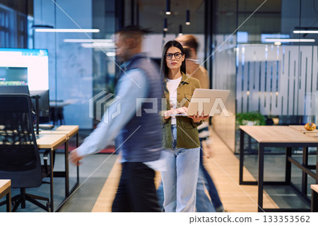 Business woman holding laptop in the middle of a dynamic modern office while colleagues move around, symbolizing focus, leadership, and productivity amid a fast-paced work environment. 133353562