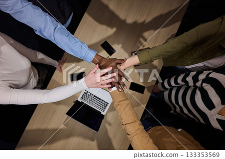 Top view of diverse coworkers business people joining hands together over a wooden office desk, symbolizing teamwork, unity, collaboration, and support in a modern workplace. 133353569
