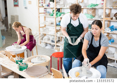 Young woman teacher helps teenagers boy and girl to make product on potter's wheel 133353572
