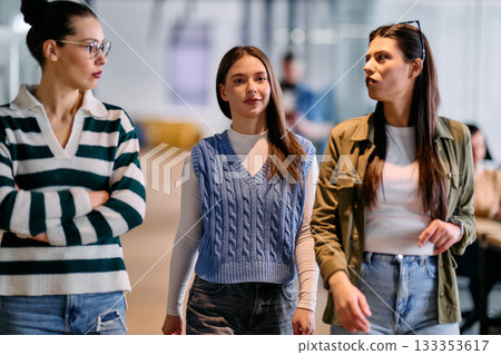 Group of three professional women walking through a modern office hallway, engaged in a casual business conversation, representing teamwork, confidence, and collaboration in the workplace. 133353617