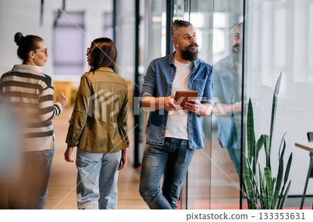 Group of coworkers walking and discussing ideas in a modern office corridor while a male colleague works at a laptop nearby, symbolizing teamwork, productivity, and creative collaboration in a 133353631