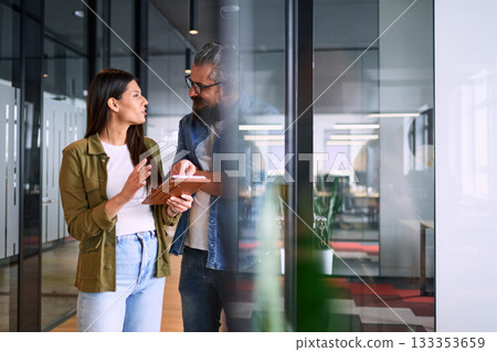 Two business professionals having a focused discussion while reviewing information on a tablet in a bright modern office hallway, symbolizing teamwork, communication, and digital collaboration. Two business professionals having a focused discussion while reviewing information on a tablet in a bright modern office hallway, symbolizing teamwork, communication, and digital collaboration. 133353659