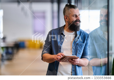 Bearded businessman standing in modern office hallway, holding digital tablet and looking confidently at camera, representing experience, leadership, and innovation in contemporary business 133353666