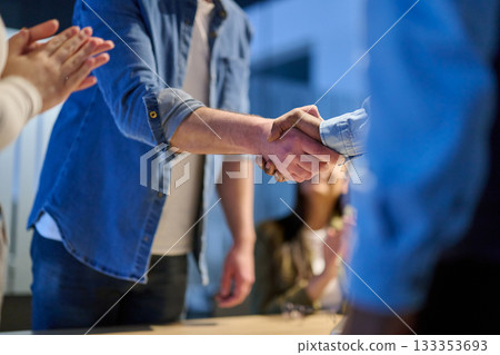 Two businessmen shaking hands after successful negotiation in a modern office, symbolizing partnership, agreement, success, and teamwork. A woman in the background applauds the deal. 133353693