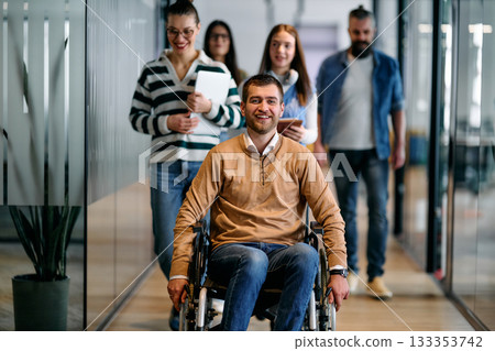 Group of diverse business people walking together in a modern office hallway, led by a confident smiling man in a wheelchair, representing inclusion, equality, and teamwork. 133353742