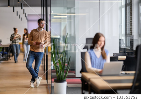 Businessman standing by glass wall in a modern office corridor, holding a digital tablet and smiling with reflection visible on the glass, symbolizing success, optimism, and modern business lifestyle. 133353752