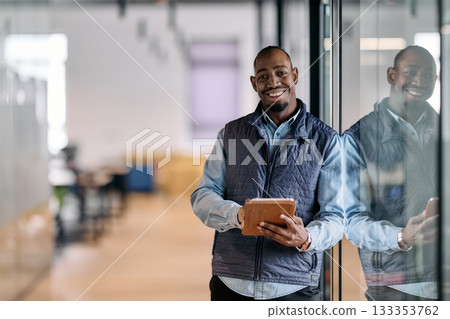 Confident african american businessman standing in a modern office hallway, smiling while using a digital tablet. Reflection in glass wall symbolizes technology, success, and innovation in business. Confident african american businessman standing in a modern office hallway, smiling while using a digital tablet. Reflection in glass wall symbolizes technology, success, and innovation in business. 133353762