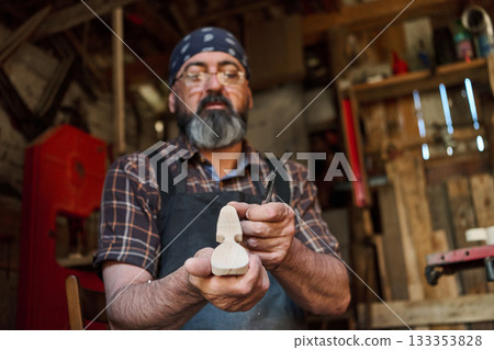 Woodcarver carefully shaping a small wooden figure in his workshop, artisan crafting a wooden sculpture, hobbyist working with hand tools, creating art in a rustic studio environment. Woodcarver carefully shaping a small wooden figure in his workshop, artisan crafting a wooden sculpture, hobbyist working with hand tools, creating art in a rustic studio environment. 133353828