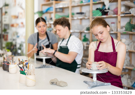 Young woman teacher helps teenagers boy and girl to sculpt ceramic product Young woman teacher helps teenagers boy and girl to sculpt ceramic product 133353829