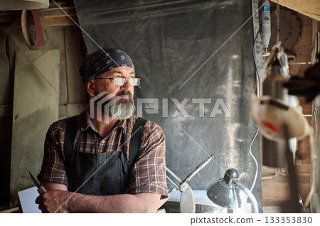 Craftsman with beard in workshop, wearing bandana and apron, holding a knife, looking thoughtful, artisan at work, small business owner, maker, entrepreneur, handmade 133353830
