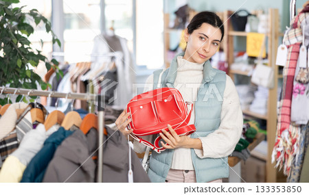 Fashionable young girl customer standing near shelves with clothes in store and choosing handbag Fashionable young girl customer standing near shelves with clothes in store and choosing handbag 133353850
