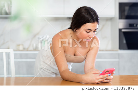 Young woman reading message on phone in kitchen 133353972