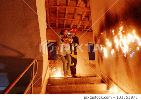 Two brave firefighters helping a frightened woman carrying her cat to safety from a burning building, symbolizing heroism, compassion, and teamwork during an emergency rescue operation. 133353973
