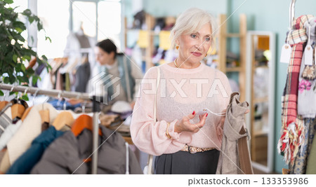 Focused happy elderly woman client choosing pants or stylish trousers in clothing mall 133353986