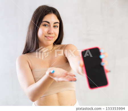 Young woman showing her mobile phone standing against wall 133353994
