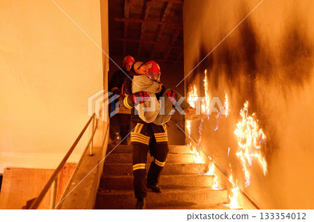 Courageous firefighter carrying a rescued woman down a burning staircase during an emergency evacuation, symbolizing bravery, heroism, and dedication to saving lives in dangerous situations. 133354012