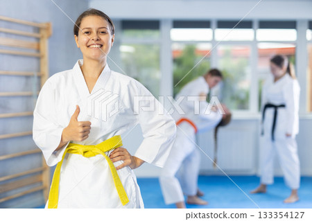 Girl in white kimono gives thumbs up and stand in gym Girl in white kimono gives thumbs up and stand in gym 133354127