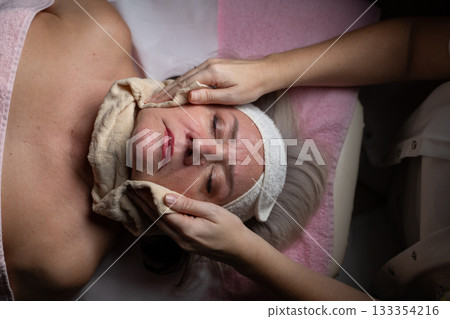 Close up of a mature woman receiving a relaxing facial massage from a professional female therapist in a modern spa. The client lies on a treatment bed with a towel and headband, enjoying skincare 133354216