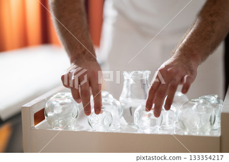 Therapist Preparing Glass Cups for Traditional Cupping Therapy. Close up of therapists hands arranging clean glass cups on a tray before starting a traditional cupping therapy session. Therapist Preparing Glass Cups for Traditional Cupping Therapy. Close up of therapists hands arranging clean glass cups on a tray before starting a traditional cupping therapy session. 133354217
