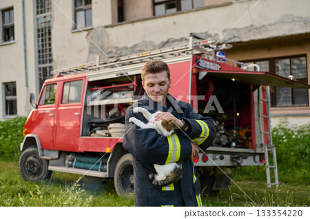 Brave firefighter holding a rescued cat in his arms near a fire truck, showing compassion and care, outdoor scene with a building in the background Brave firefighter holding a rescued cat in his arms near a fire truck, showing compassion and care, outdoor scene with a building in the background 133354220
